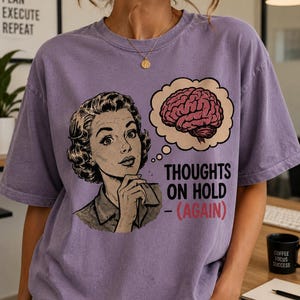 Puede incluir: Una camiseta morada de manga corta con una ilustración gráfica de una persona y una burbuja de pensamiento que contiene un cerebro. El texto dice THOUGHTS ON HOLD - (AGAIN) en letras negras y rojas. La camiseta tiene cuello redondo y un corte holgado.