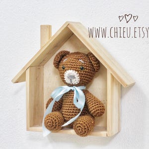 May include: A handmade brown crochet teddy bear with a light blue ribbon, displayed inside a wooden house-shaped shelf. The bear has a white muzzle and is set against a white wall. The website address www.chieu.etsy.com is visible.