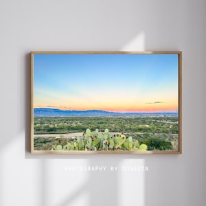 May include: A framed landscape photograph featuring a desert scene with a vibrant sunset. The foreground shows a prickly pear cactus with red fruit. The middle ground has green trees and a city. The background has mountains under a blue and orange sky. The text "PHOTOGRAPHY BY DONAVIN" is at the bottom.