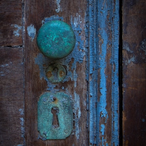 May include: A close-up of a weathered wooden door with a teal-colored door knob and keyhole. The door is painted blue, but the paint is peeling and chipping.