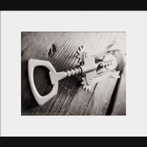 May include: A silver corkscrew with a metal handle and a worm screw lies on a wooden surface. The corkscrew is in focus, while the wooden surface is blurred in the background.