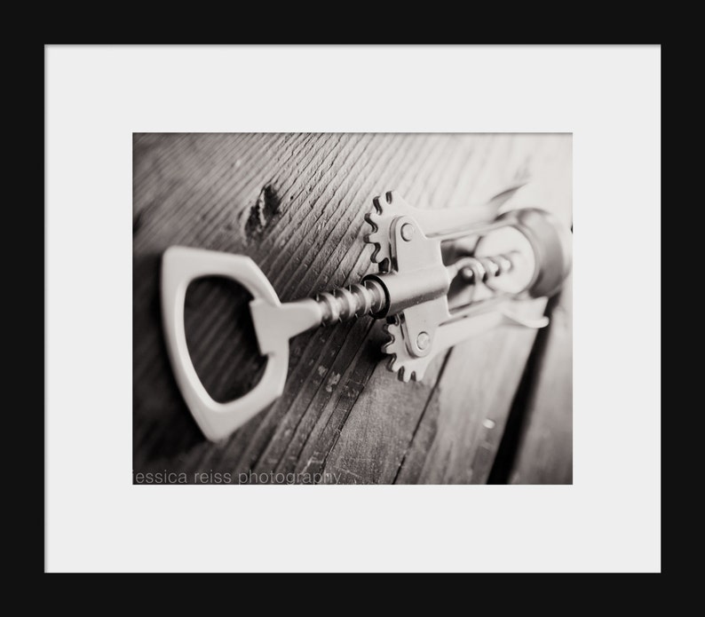 May include: A silver corkscrew with a metal handle and a worm screw lies on a wooden surface. The corkscrew is in focus, while the wooden surface is blurred in the background.