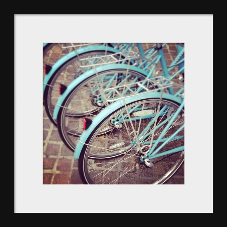 May include: A row of four blue bicycles parked on a brick pathway. The bicycles have silver spokes and silver handlebars. The front wheels are in focus, and the rear wheels are out of focus.