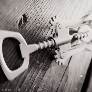 May include: A silver metal bottle opener with a corkscrew and a lever. The opener is lying on a wooden surface.