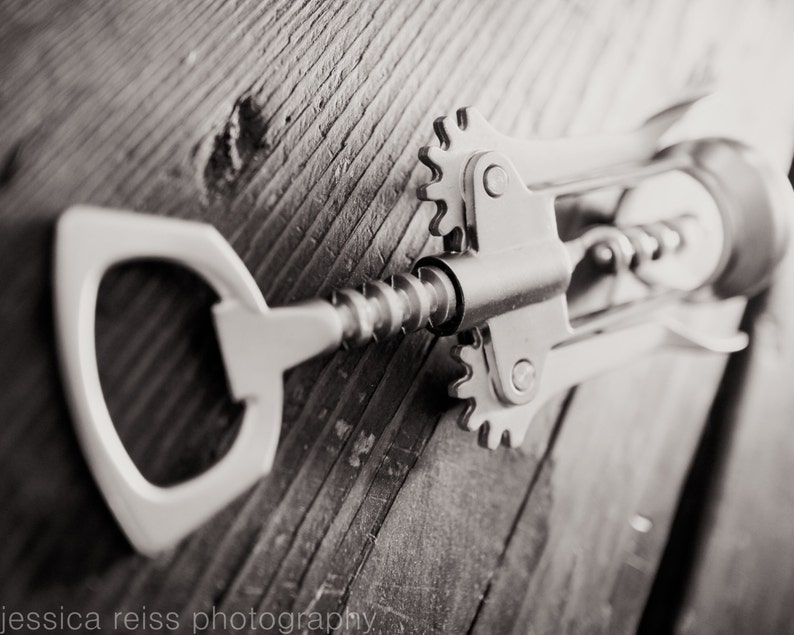 May include: A silver metal bottle opener with a corkscrew and a lever. The opener is lying on a wooden surface.