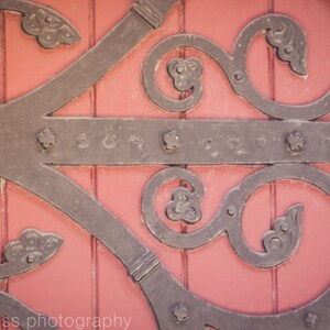 May include: A close-up of a decorative metal door hinge with intricate scrollwork and a red wooden background.