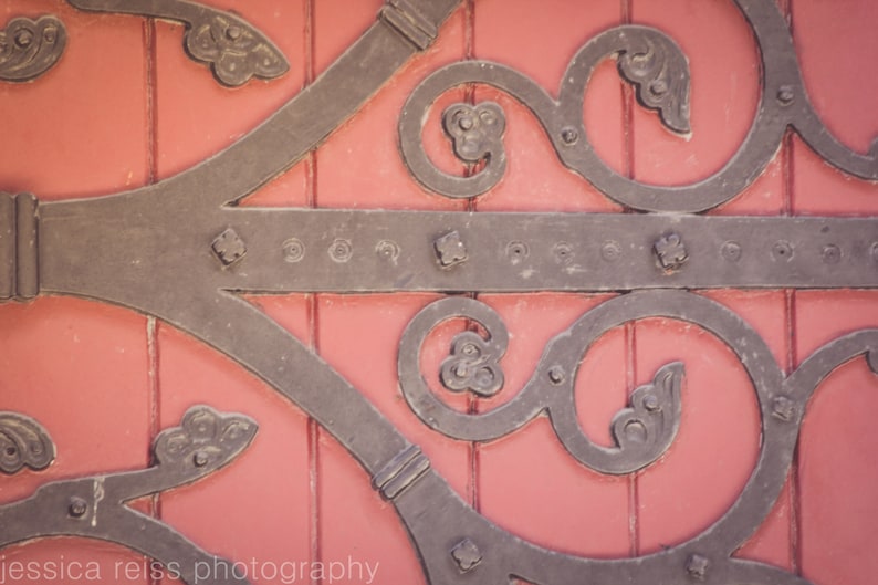 May include: A close-up of a decorative metal door hinge with intricate scrollwork and a red wooden background.