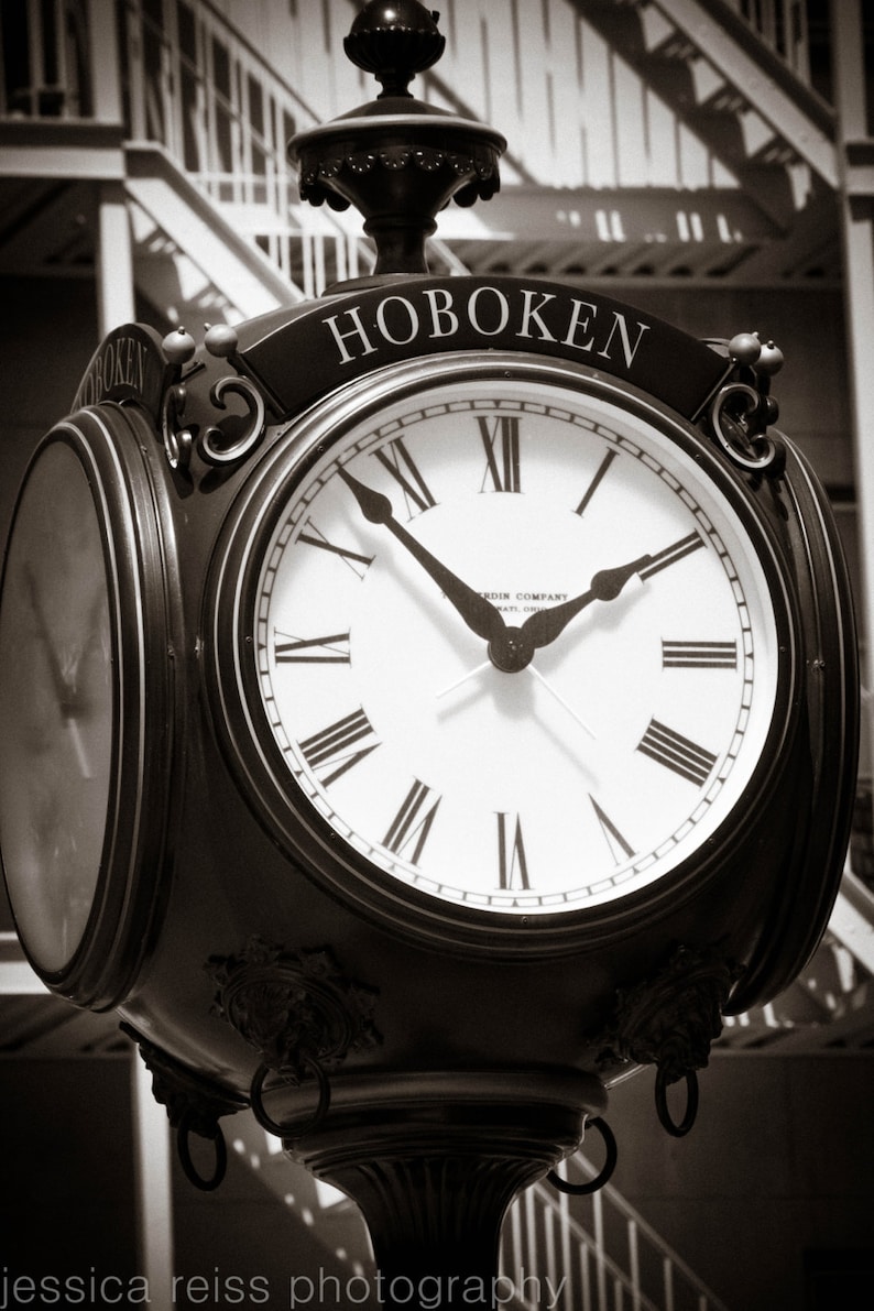 Op de afbeelding: Een zwarte klok met Romeinse cijfers en het woord "HOBOKEN" op de wijzerplaat. De klok is gemonteerd op een paal en heeft een decoratief ontwerp.
