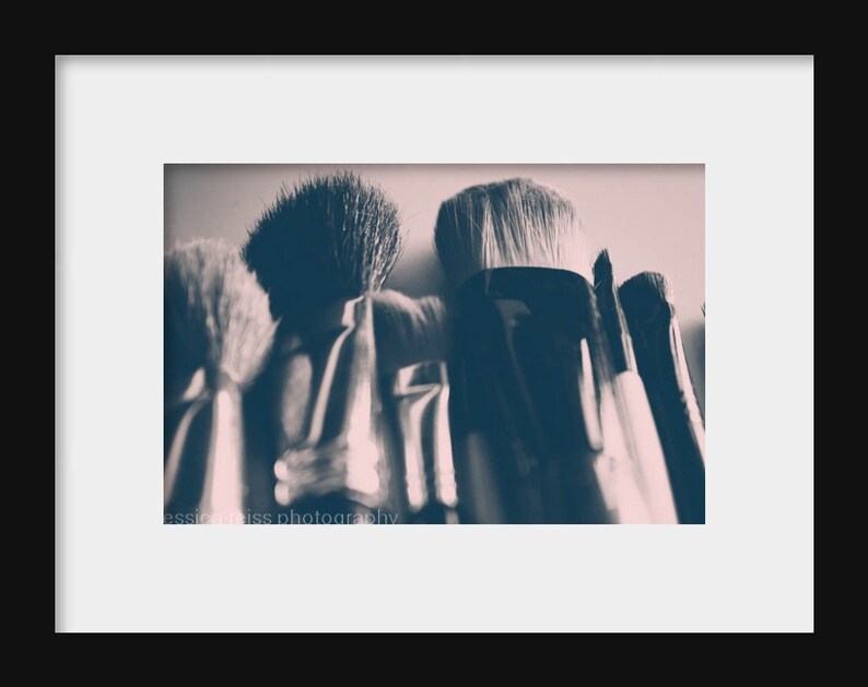 May include: Close-up of a collection of makeup brushes with dark handles and soft bristles. The brushes are arranged closely together, with the bristles facing upwards. The image has a soft, muted color palette, with a focus on texture and detail. The text "jessica weiss photography" is visible.