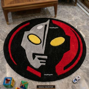 Pu&ograve; includere: Un tappeto rotondo nero con un design di personaggio stilizzato in grigio, giallo e rosso. Il design include il testo "SatyaRugsArt" e "KAUU HUNTER". Il tappeto &egrave; posto su un pavimento effetto marmo.