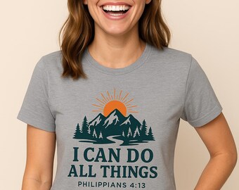 Può includere: T-shirt grigio con grafica di montagna e sole. Il design include il testo "I CAN DO ALL THINGS" e "PHILIPPIANS 4:13". La maglietta è a maniche corte e ha un girocollo.