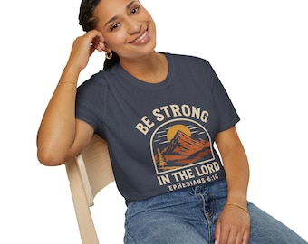 May include: Navy blue t-shirt with a mountain and sun graphic. The text on the shirt reads "BE STRONG IN THE LORD" and "Ephesians 6:10". The design is in shades of orange, brown, and beige.
