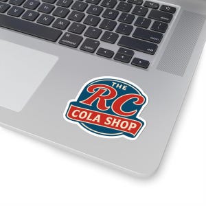 May include: A sticker featuring the text "THE RC COLA SHOP" in a retro design. The sticker has a blue background with red and white accents, and is placed on a laptop.