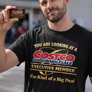 Puede incluir: Camiseta negra con el logo de Costco Wholesale y el texto "YOU ARE LOOKING AT A COSTCO EXECUTIVE MEMBER I'm Kind of a Big Deal." Una persona sostiene una tarjeta de miembro ejecutivo de Costco.
