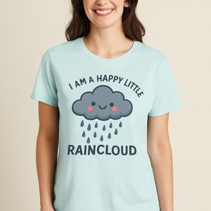 May include: Pale blue t-shirt featuring the text "I AM A HAPPY LITTLE RAINCLOUD" and a cartoon cloud design. The cloud is grey with a smiling face and pink cheeks, with blue raindrops falling.