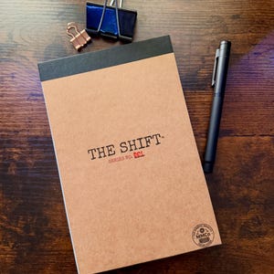May include: A tan notepad with a black top binding, featuring the text THE SHIFT and SERIES NO. 001 in black and red ink. A black pen lies to the right, while a large silver binder clip and a small copper binder clip sit above the notepad.