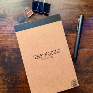 May include: A tan notepad with a black top binding, labeled THE FOCUS SERIES NO. 001 in black and red text, accompanied by a black metal pen, a large black binder clip, and a small copper-colored binder clip, featuring a circular logo in the corner.