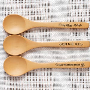 May include: Three wooden spoons with engraved text. The top spoon reads "My Kitchen My Rules", the middle spoon reads "Made With Love", and the bottom spoon reads "Bake the Season Bright". Each spoon has a rounded bowl and a long handle.