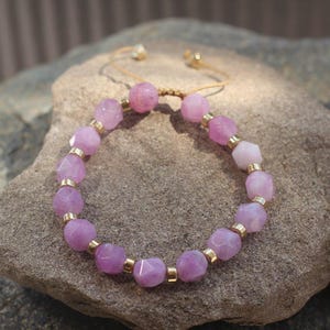 Op de afbeelding: Een armband met facetgeslepen, lichtpaarse edelstenen en goudkleurige metalen accenten. De armband is geregen op een beige koord, met verstelbare goudkleurige kralen aan de uiteinden. De armband wordt gepresenteerd op een grijs stenen oppervlak.