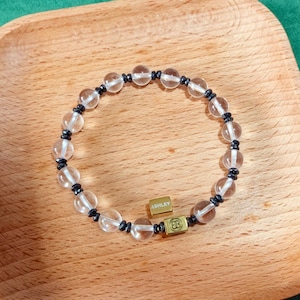 Puede incluir: Una pulsera de cuentas con una alternancia de cuentas esféricas transparentes y pequeñas cuentas de disco metálicas oscuras. La pulsera incluye dos dijes rectangulares dorados, uno grabado con el nombre ASHLEY y el otro con un símbolo.