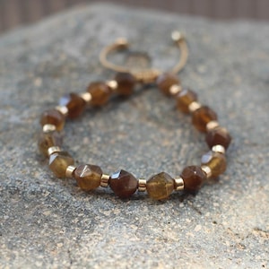 Puede incluir: Una pulsera con piedras preciosas facetadas de color ámbar y pequeñas cuentas doradas. La pulsera ajustable tiene un cordón tejido. Las piedras preciosas se muestran sobre una superficie gris texturizada.