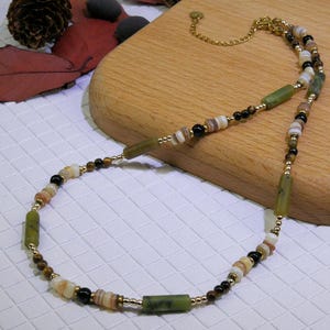 Puede incluir: Un collar artesanal con una mezcla de cuentas verdes, marrones, negras y blancas. El collar incluye cuentas rectangulares verdes, pequeñas cuentas redondas y eslabones de cadena dorados. El collar se muestra sobre una superficie de madera.