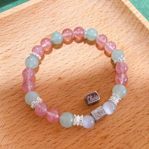 Puede incluir: Una pulsera de cuentas con cuentas redondas alternas de color rosa y verde pálido. La pulsera incluye espaciadores de tono plateado y dos dijes cuadrados de tono plateado, uno grabado con el nombre Olivia y el otro con un símbolo.