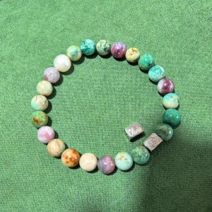 May include: A beaded bracelet featuring a variety of round, multi-colored stones in shades of green, pink, cream, and white. The bracelet includes two silver-toned metal charms, one of which is rectangular and engraved with the name Olivia.