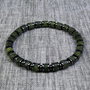 May include: A green and black beaded bracelet. The bracelet features round, olive-green beads interspersed with dark grey metallic spacers. The beads are strung together to form a circular shape, suitable for wrist wear. The bracelet is displayed on a textured grey surface.