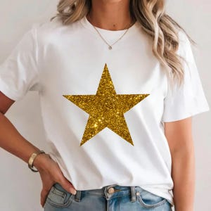 May include: White t-shirt with a large, gold glitter star. The star is a five-pointed shape, centrally placed on the front. The shirt has short sleeves and a round neck, a classic casual style.