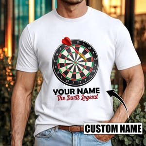 Op de afbeelding: Wit T-shirt met een dartbord ontwerp en een rode dartpijl. Het shirt toont de tekst "YOUR NAME The Darts Legend" en een "CUSTOM NAME" vak. Het dartbord heeft zwarte, rode, groene en witte details.