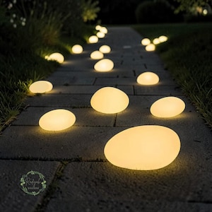 May include: A stone pathway illuminated by glowing, oval-shaped outdoor lights, resembling smooth stones. The lights are arranged along a stone walkway, creating a warm, inviting ambiance. The scene is set outdoors, with grass and foliage visible.