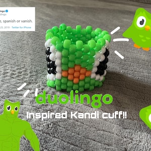 May include: A green, black, white, and orange Kandi cuff inspired by Duolingo. The cuff is made of small beads and features the Duolingo owl mascot. The image also includes the text "It's simple, spanish or vanish."