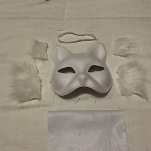 May include: A white cat mask with eye cutouts, a nose, and ear shapes. The mask is surrounded by white faux fur pieces, a white elastic band, and a white felt square. A DIY craft project.