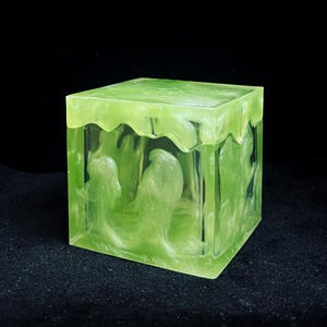 May include: A translucent, lime green cube-shaped box with a wavy top edge. The interior features a swirling, lighter green design. The box is set against a black background, highlighting its unique, artistic design.