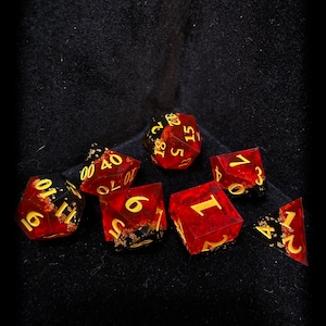 May include: A collection of seven red and gold dice, each with gold numbering. The dice are various shapes, including a six-sided die, a ten-sided die, and a twenty-sided die. The dice are scattered on a dark, textured surface.