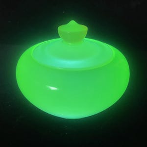 May include: A glowing green glass jar with a lid. The jar is round with a slightly flattened base. The lid is a lighter shade of green and has a small, pointed knob on top. The jar is set against a black background.