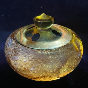 May include: A small, round, amber-colored glass jar with a textured surface. The jar has a gold-colored metal lid with decorative cutouts and a small, rounded finial. The jar is likely used for storing small items or as a decorative piece.