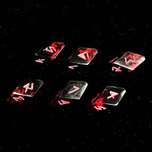 May include: Six rectangular, silver-colored rune stones with red accents and symbols. The runes are arranged on a black surface, with each stone displaying a unique symbol. The stones appear to be made of a reflective material.