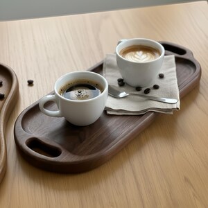 May include: Two white coffee cups filled with coffee and latte art sit on a dark brown wooden serving tray. A small silver spoon and a linen napkin are also on the tray, along with coffee beans. The tray has a handle cutout.