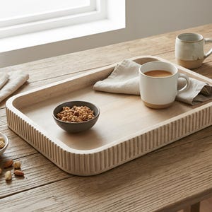 Peut inclure: Un plateau de service en bois clair avec un bord cannelé contient une tasse blanche avec du café, un petit bol de granola et une serviette en lin pliée. Le plateau est posé sur une table en bois près d'une fenêtre.
