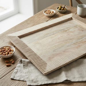 Könnte beinhalten: Ein rechteckiges Serviertablett aus hellem Holz mit einer strukturierten, geriffelten Umrandung. Das Tablett ist leer und steht auf einem Holztisch mit kleinen Schalen mit Nüssen und Oliven.