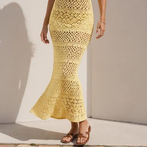 May include: A floor-length, pale yellow crochet dress featuring a halter neckline and a flared, mermaid-style hem. The garment is constructed with various intricate, open-work stitch patterns throughout the bodice and skirt, creating a textured, semi-sheer appearance that extends down to the ankles.