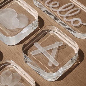 Pode incluir: Tábua de cortar acrílica transparente com vários desenhos. Uma tábua de cortar tem a palavra "hello" em uma fonte de escrita. Outras tábuas de cortar apresentam desenhos de coração, flor e jogo da velha. As tábuas de cortar estão sobre uma superfície de madeira.