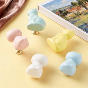 May include: A collection of six cat-shaped cabinet knobs in pastel colors: light blue, pink, yellow, white, and blue. Each knob is mounted on a gold-colored base. The knobs are arranged on a yellow surface.