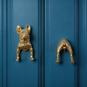 May include: Gold-colored cabinet hardware featuring a French Bulldog design. One handle depicts the dog's head, the other its rear. The hardware is mounted on a blue cabinet door, adding a unique and playful touch to home decor.