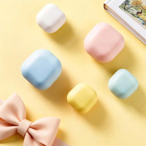 Op de afbeelding: Een bovenaanzicht toont zes pastelkleurige, vierkante objecten met afgeronde hoeken, gerangschikt op een gele ondergrond. De kleuren zijn wit, roze, blauw, geel en lichtblauw. Een roze strik en een boek zijn ook zichtbaar.