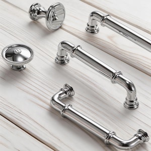 May include: Close-up of various polished silver cabinet hardware, including knobs and handles. The hardware has a classic design with decorative detailing. The pieces are arranged on a light-colored wooden surface, showcasing their reflective qualities.