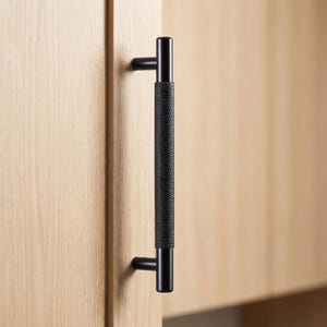 May include: Black cabinet pull with a textured center grip and smooth, rounded ends. The handle is mounted on a light-colored wooden cabinet door. The hardware has a modern, minimalist design.