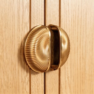 May include: Close-up of a gold-colored cabinet pull with a half-circle design and a ribbed texture. The pull is mounted on a light-colored wooden cabinet door with vertical grain.